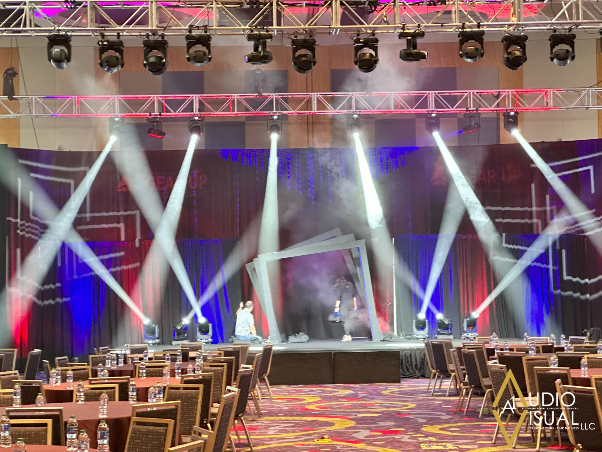 Ballroom general session stage with truss, moving-head beam lights and scenic frame set over banquet rounds